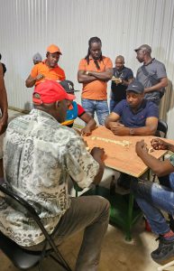 A group of men sitting around a wooden table playing dominos with a group of onlookers behind them